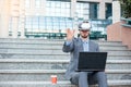 Successful young businessman using VR goggles and making hand gestures, working on a laptop in front of an office building Royalty Free Stock Photo