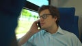 Handsome young businessman wearing a blue shirt and glasses is riding a train dialing a number and talking on his phone