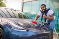 Handsome young casual African American man in t-shirt and jeans wiping modern blue electric car hood with red microfiber Royalty Free Stock Photo