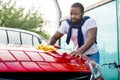 Handsome young casual African American man in t-shirt and jeans wiping red car hood with yellow microfiber cloth at the Royalty Free Stock Photo