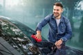 Handsome young casual Caucasian man in jeans shirt wiping his modern car windshield with red microfiber cloth at the Royalty Free Stock Photo