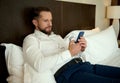 Handsome young Caucasian bearded man, successful businessman browsing and chatting on mobile phone, lying on a bed in the Royalty Free Stock Photo