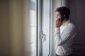 Handsome young caucasian man standing in white shirt, looking out the window, talking on his cell phone Royalty Free Stock Photo