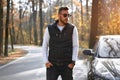 A handsome young Caucasian man wearing style black vest standing near black car on nature Royalty Free Stock Photo