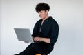 Handsome young curly guy in total black look sitting on chair and playing computer games on modern laptop, gray studio Royalty Free Stock Photo