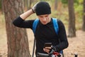 Handsome young European cyclist wearing black sportwear and blue bacpack, messaging friends on mobile phone after lost in woods,