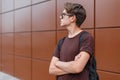 Handsome young fashionable hipster man in stylish sunglasses in a brown t-shirt with a backpack poses near a modern building Royalty Free Stock Photo