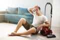 Handsome young fatigued man sitting near vacuum cleaner in the living room looking tired after cleaning appartment house Royalty Free Stock Photo