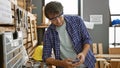 Handsome young guy with glasses, hispanic carpenter texting on his smartphone in a cozy carpentry workshop