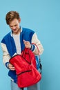 a handsome young happy man in stylish clothes puts a notebook in his red backpack while standing on a light blue Royalty Free Stock Photo