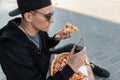 Handsome young hipster fashionable man in stylish cap in sunglasses in black sweatshirt eats pizza sitting on a tile in the city. Royalty Free Stock Photo