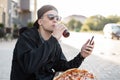 Handsome young hipster man in a black cap in sunglasses hoodie sits on the street with a phone in his hands with a pizza Royalty Free Stock Photo