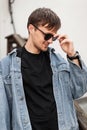 Handsome young hipster man with a cute smile straightens sunglasses outdoors. Attractive guy in a fashionable denim jacket Royalty Free Stock Photo