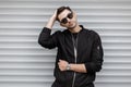 Handsome young hipster man in a fashionable black jacket in stylish sunglasses is posing outdoors near a white metal modern wall. Royalty Free Stock Photo