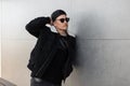 Handsome young hipster man in a fashionable jacket with white fur in sunglasses in a black stylish cap in a black T-shirt in jeans Royalty Free Stock Photo