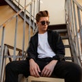 Handsome young hipster man with hairstyle in trendy clothes in stylish sunglasses poses sitting Royalty Free Stock Photo