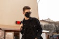 Handsome young hipster man with hairstyle in trendy sunglasses in fashionable black denim jacket in stylish medical black mask Royalty Free Stock Photo