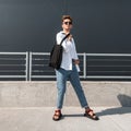 Handsome young hipster man in red sandals in white shirt in sunglasses in vintage jeans with a cloth bag is enjoying the sun Royalty Free Stock Photo