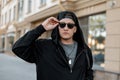 Handsome young hipster man in stylish black clothes in a trendy cap in dark sunglasses walks around the city on a summer day. Royalty Free Stock Photo