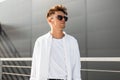 Handsome young hipster man with stylish hairstyle in black sunglasses in trendy white shirt in T-shirt poses near a gray building Royalty Free Stock Photo