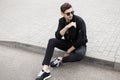 Handsome young hipster man in a stylish jacket in black jeans in sneakers in trendy sunglasses is sitting on the sidewalk Royalty Free Stock Photo