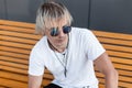 Handsome young hipster man in trendy white t-shirt in stylish blue sunglasses with amulets on a neck is sitting on a wooden bench Royalty Free Stock Photo