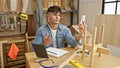 Handsome young hispanic carpenter expertly managing business via video call from his buzzing indoor carpentry workshop, surrounded
