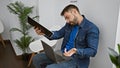 Handsome young hispanic man multitasking with laptop, clipboard and phone call in waiting room