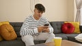 Handsome young hispanic man relaxing at home, using his smartphone on sofa, immersed in technology, enjoying coffee, showing Royalty Free Stock Photo