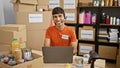 Handsome young hispanic man selflessly volunteering, working on laptop with headphones at charity center, donations box nearby Royalty Free Stock Photo