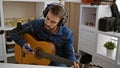 Handsome young hispanic man sitting in a music studio, passionately playing a chord on a classical guitar while looking at his Royalty Free Stock Photo