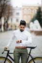 Handsome young indian man standing with his bicycle on the street in city center and looking on watch Royalty Free Stock Photo