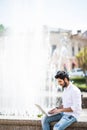 Handsome young indian man using laptop in city near fountain on a summer day. Royalty Free Stock Photo