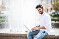 Handsome young indian man using laptop in city near fountain on a summer day. Royalty Free Stock Photo