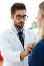 Handsome young male doctor checking beautiful young woman patient heartbeat using stethoscope in medical office. Royalty Free Stock Photo