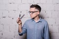 Handsome young male hairdresser in glasses, posing with scissors and comb, on gray brick wall background, horizontal Royalty Free Stock Photo