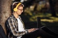 Handsome young male student in casual outfit with yellow headphones using notebook or laptop while sitting on the grass at the Royalty Free Stock Photo