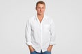 Handsome young male in white shirt, keeps hands in pockets, looks directly at camera, has serious confident expression, isolated o Royalty Free Stock Photo