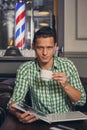 Handsome young man in a barbershop is waiting for the master in the waiting room while drinking coffee