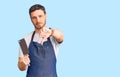 Handsome young man with bear wearing professional apron holding knife with angry face, negative sign showing dislike with thumbs Royalty Free Stock Photo