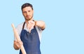 Handsome young man with bear wearing professional baker apron holding kneading roll with angry face, negative sign showing dislike
