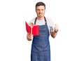 Handsome young man with bear wearing professional baker apron reading cooking recipe book annoyed and frustrated shouting with Royalty Free Stock Photo