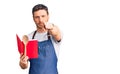 Handsome young man with bear wearing professional baker apron reading cooking recipe book pointing with finger to the camera and Royalty Free Stock Photo