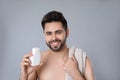 Handsome young man with beard holding post lotion on grey background
