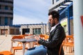Handsome young man with beard, sculptural body, is sitting in a bar while looking at his mobile phone. He is in a square in a Royalty Free Stock Photo