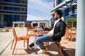 handsome young man with beard, sculptural body, is sitting in a bar while looking at his mobile phone. He is in a square in a Royalty Free Stock Photo