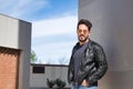 Handsome young man with beard, sunglasses, leather jacket and jeans, with his hands in his pockets and looking at the camera. Royalty Free Stock Photo