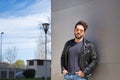 Handsome young man with beard, sunglasses, leather jacket and jeans, leaning against the wall of a building, hands in his pockets Royalty Free Stock Photo