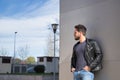 Handsome young man with beard, sunglasses, leather jacket and jeans, leaning against the wall of a building, hands in his pockets Royalty Free Stock Photo