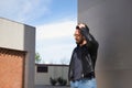 Handsome young man with beard, sunglasses, leather jacket and jeans, leaning against the wall of a building touching his hair. Royalty Free Stock Photo